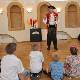 RATATATA SP. Z O.O. - Iluzjonista w stroju scenicznym prezentuje triki grupie dzieci siedzących na podłodze. W tle rekwizyty i dekoracje sali.