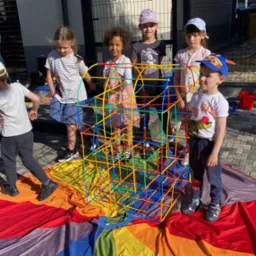 Grupa dzieci buduje kolorową konstrukcję z rurek na tęczowej płachcie na zewnątrz, w tle budynek.