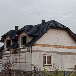 JBDOM Jarosław Brzyś - Surowy stan domu z białych bloczków z zamontowanym czarnym dachem z blachodachówki, widoczne rusztowania i drewniane wzmocnienia wzdłuż ścian.