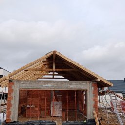 JBDOM Jarosław Brzyś - Budowa domu z czerwonej cegły w trakcie wznoszenia dachu; widoczne drewniane elementy konstrukcyjne i fragmenty innych budynków w tle.