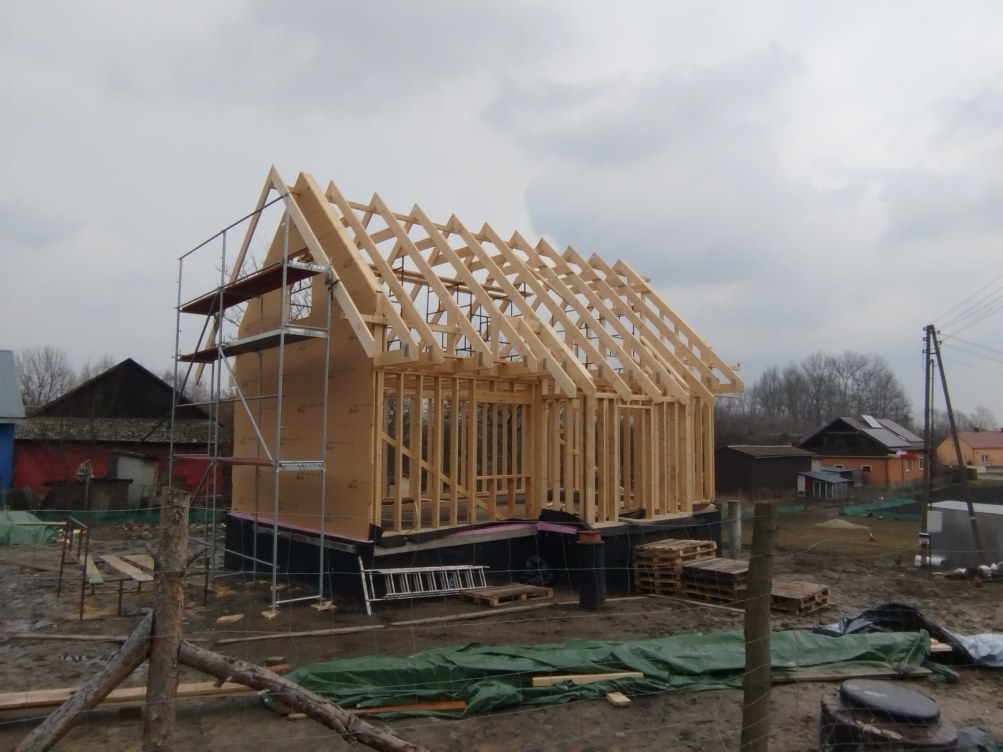 Wznoszenie konstrukcji drewnianej domu szkieletowego, widoczne rusztowanie, elementy więźby dachowej, ściany szkieletowe, teren budowy w trakcie prac.