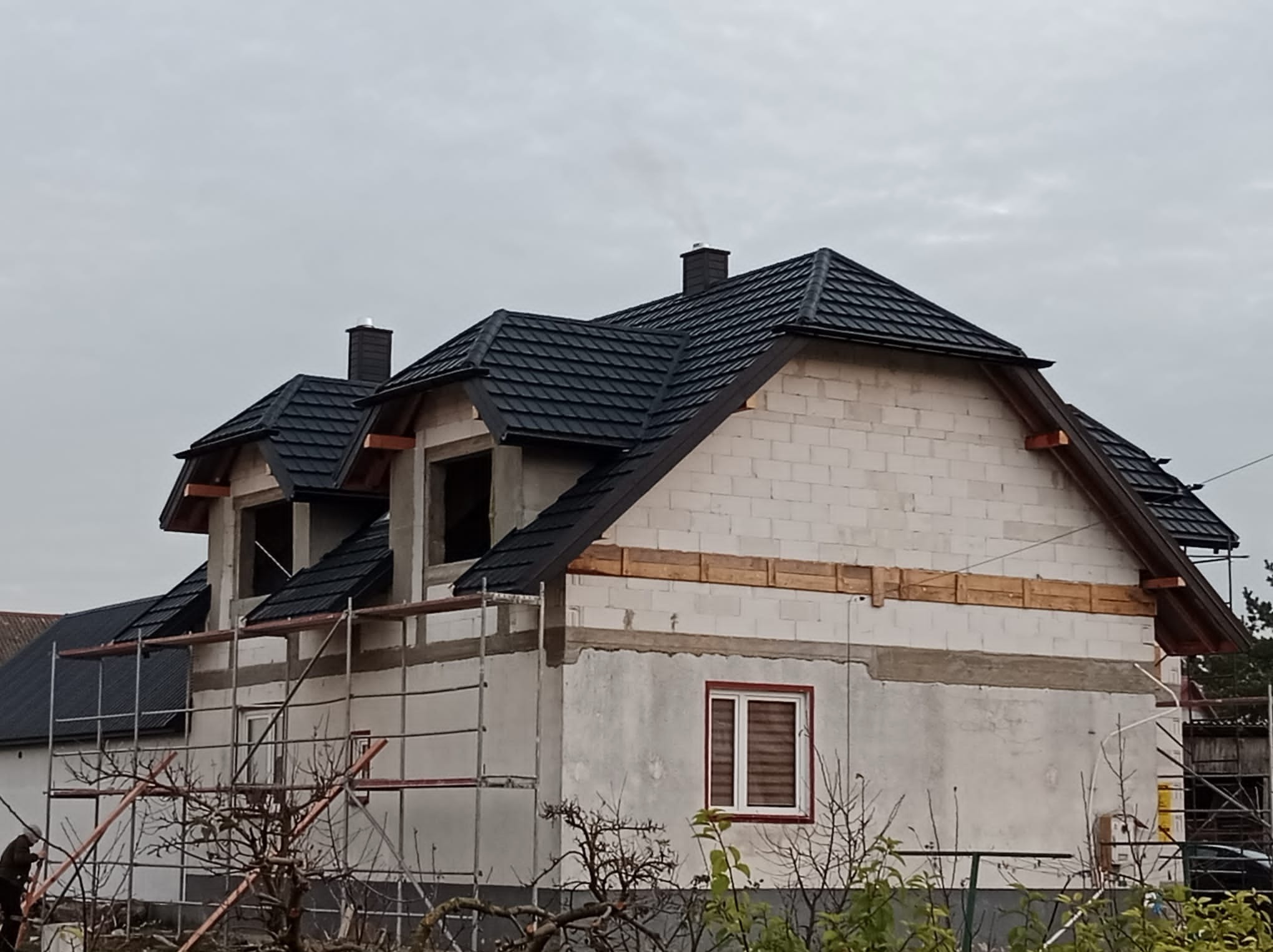 Surowy stan domu z białych bloczków z zamontowanym czarnym dachem z blachodachówki, widoczne rusztowania i drewniane wzmocnienia wzdłuż ścian.
