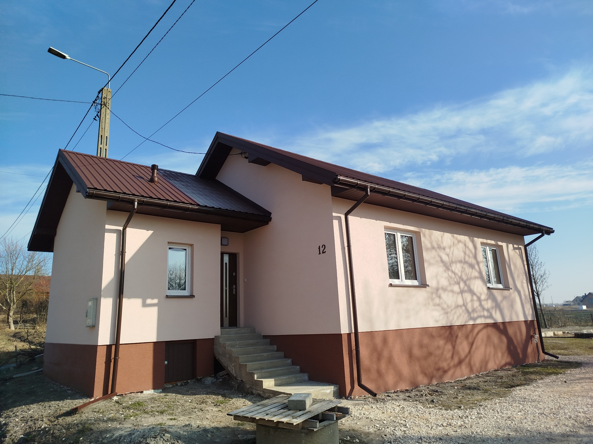 Niski, parterowy dom z brązowym dachem i jasnoróżową elewacją, z widocznym numerem '12' nad schodami wejściowymi, na tle błękitnego nieba z chmurami.