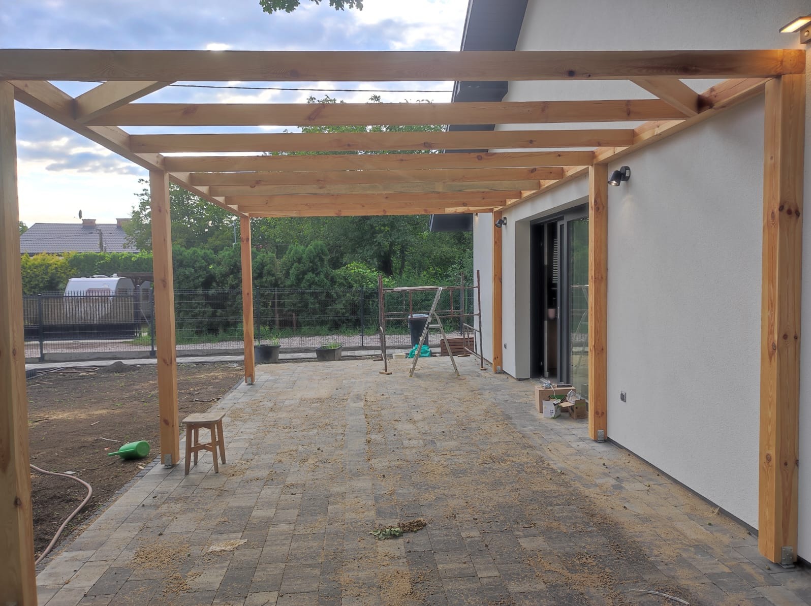 Drewniana pergola w trakcie budowy, widoczne słupy i belkowanie, na ziemi kostka brukowa z resztkami trocin, drabina oparta o ścianę budynku.