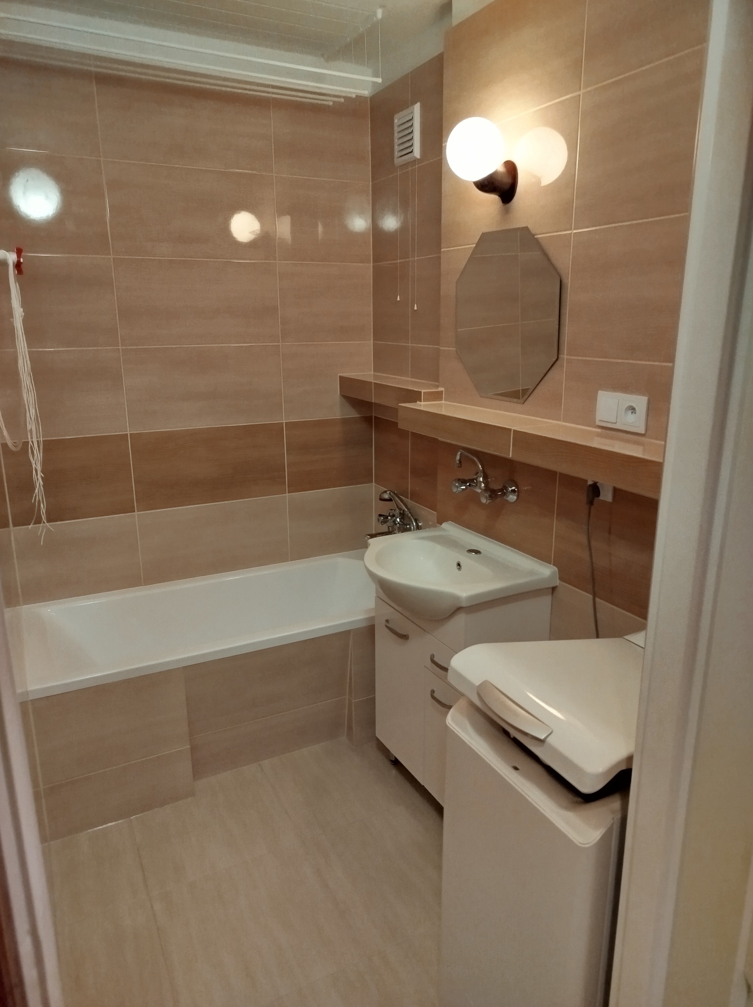 Wykończona łazienka z beżowymi płytkami, białą wanną i umywalką z szafką, ośmiokątnym lustrem i pralką ładowaną od góry.