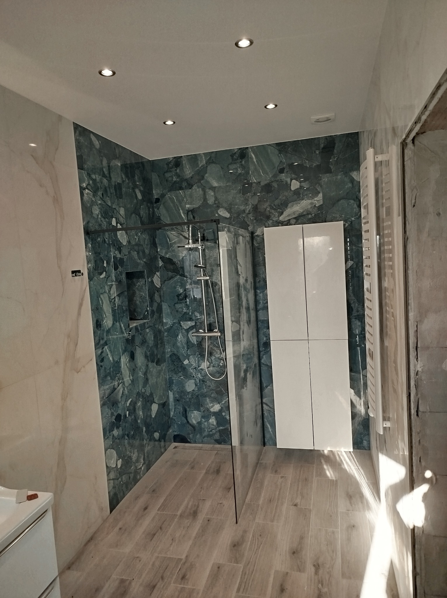 Nowoczesna łazienka z kabiną prysznicową typu walk-in, wykończona płytkami imitującymi marmur w odcieniach zieleni i bieli, drewnopodobna podłoga, biała szafka łazienkowa i grzejnik drabinkowy.