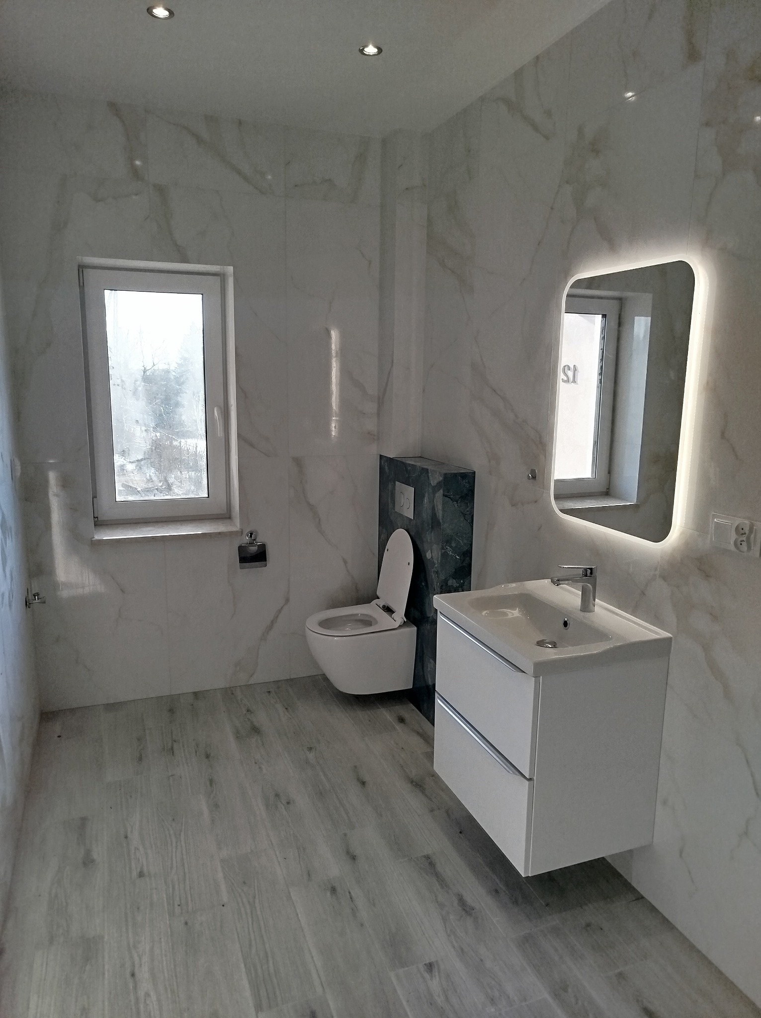 Wykończona łazienka w jasnych marmurowych płytkach z drewnopodobną podłogą, wisząca toaleta z zabudową w ciemnym odcieniu zieleni, biała szafka pod umywalkę z podświetlanym lustrem i oknem.