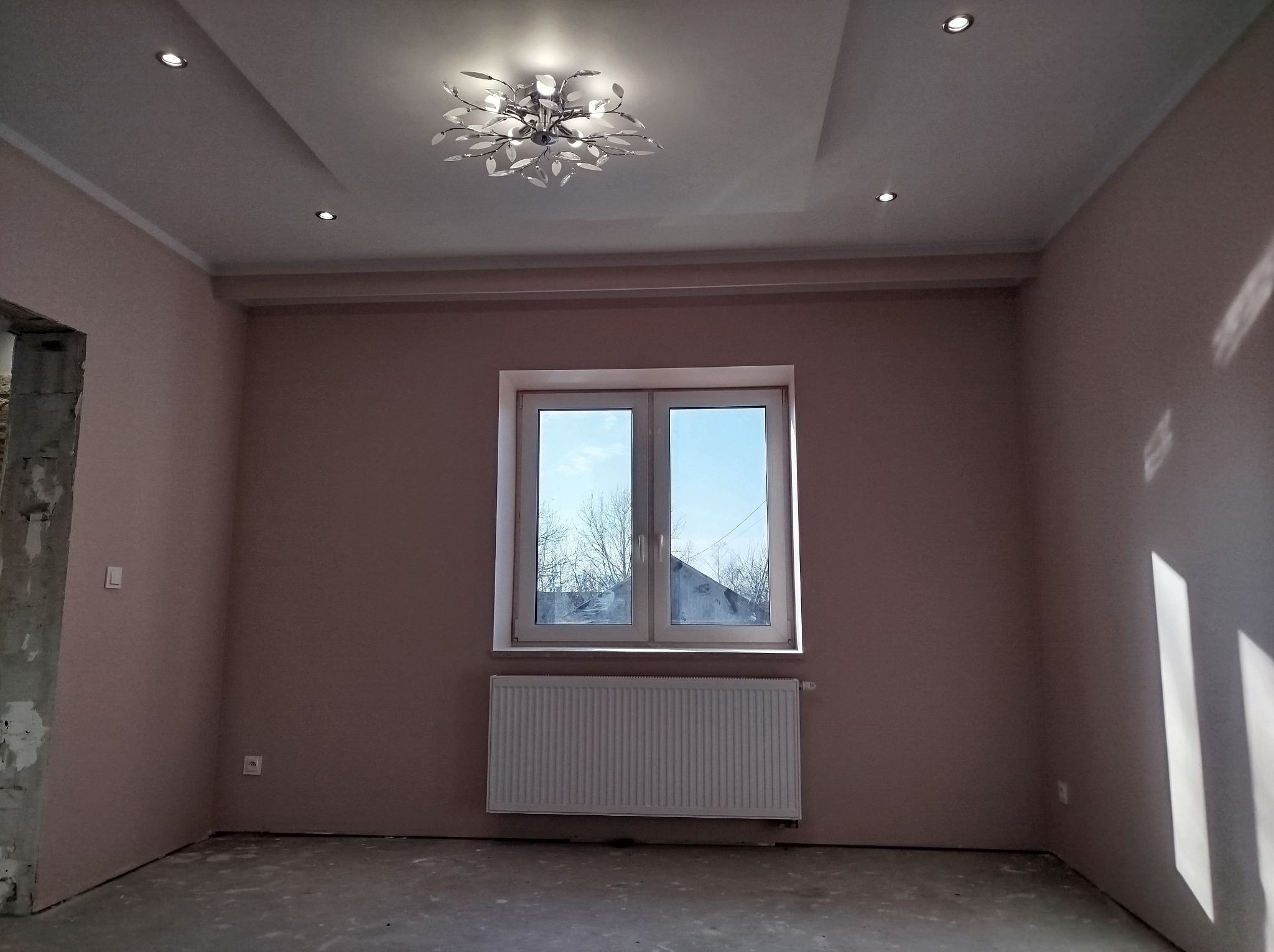 Wnętrze pokoju w trakcie wykańczania: pomalowane ściany w odcieniach beżu, białe okno z grzejnikiem, sufit z ozdobnym żyrandolem w kształcie liści i punktowymi lampami, widoczny fragment ściany...