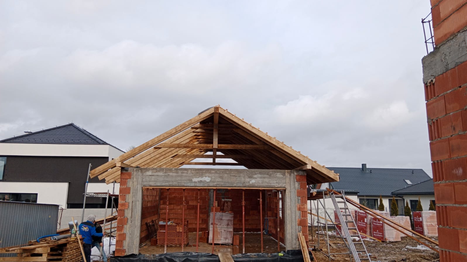 Budowa domu z czerwonej cegły w trakcie wznoszenia dachu; widoczne drewniane elementy konstrukcyjne i fragmenty innych budynków w tle.