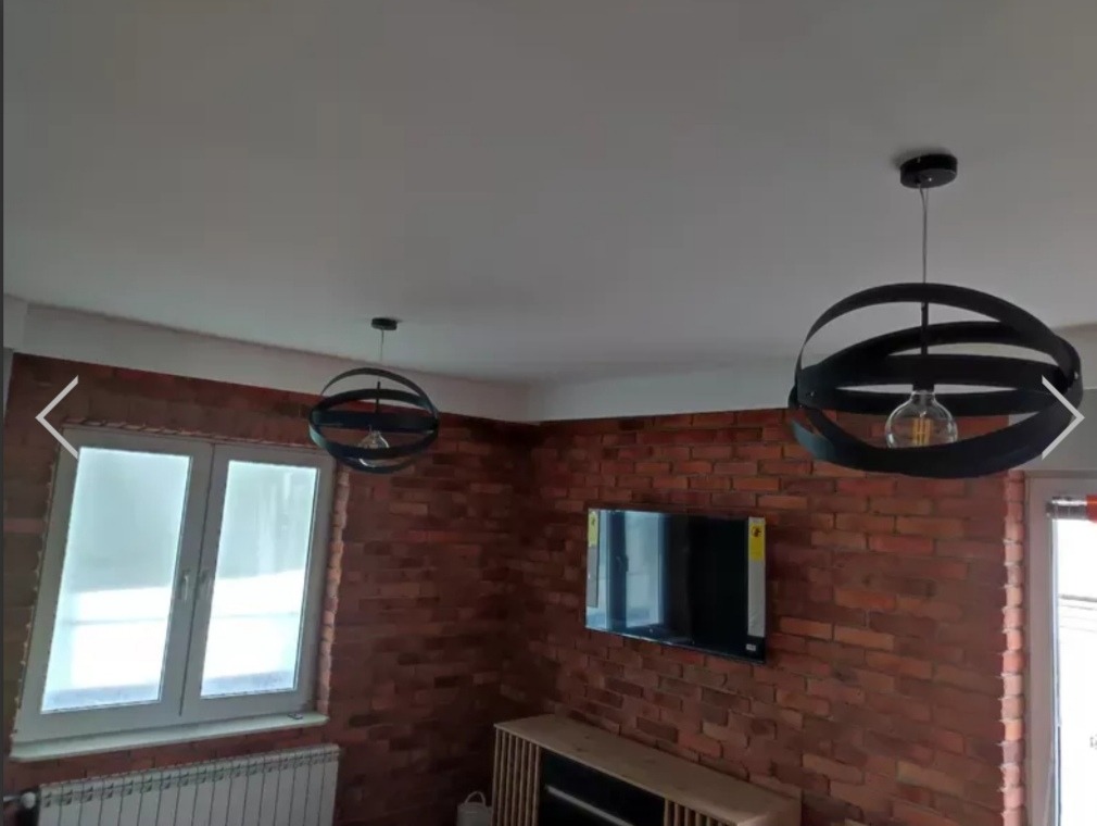 Wnętrze pokoju z ceglaną ścianą w Busko-Zdroju, oświetlone dwoma nowoczesnymi, czarnymi lampami wiszącymi z żarówkami Edisona, widoczny telewizor i białe okno z grzejnikiem.