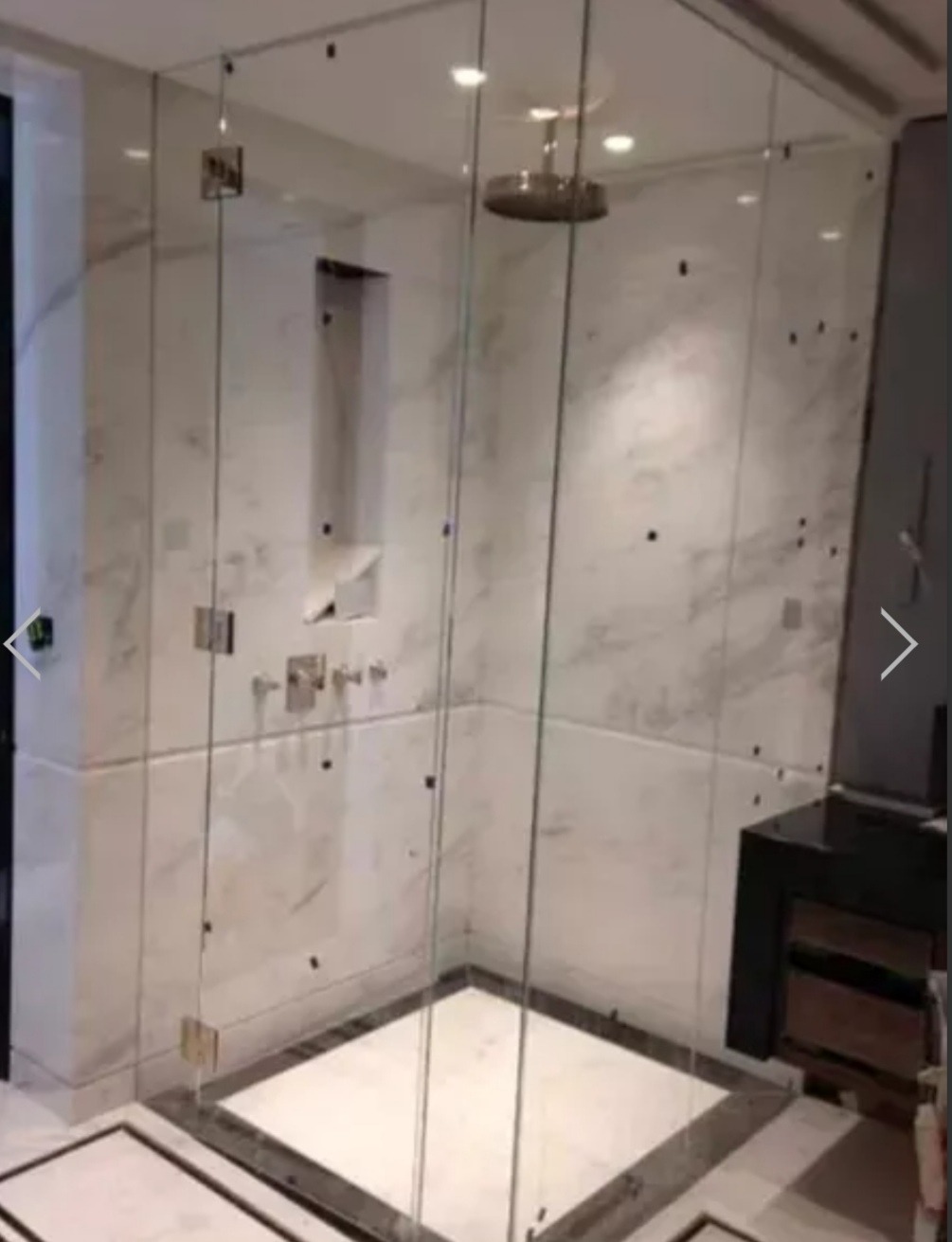 Nowoczesna kabina prysznicowa ze szklanymi ścianami i marmurowymi płytami, widoczne chromowane elementy armatury i deszczownica sufitowa.