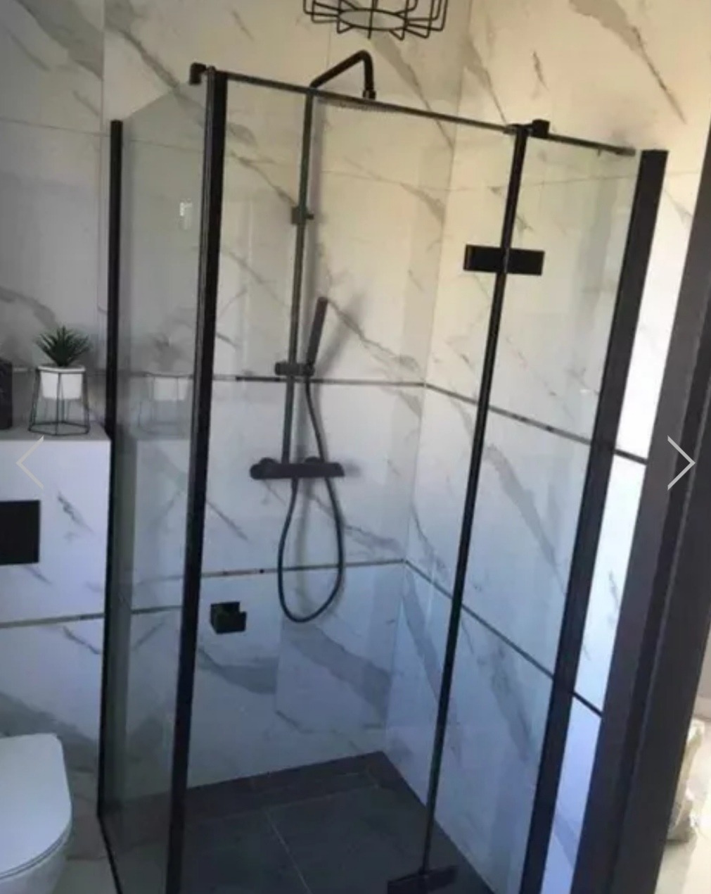 Nowoczesna kabina prysznicowa z czarnymi profilami i deszczownicą, na tle białych marmurowych płytek, z widocznym fragmentem toalety i szafki z dekoracyjną rośliną.