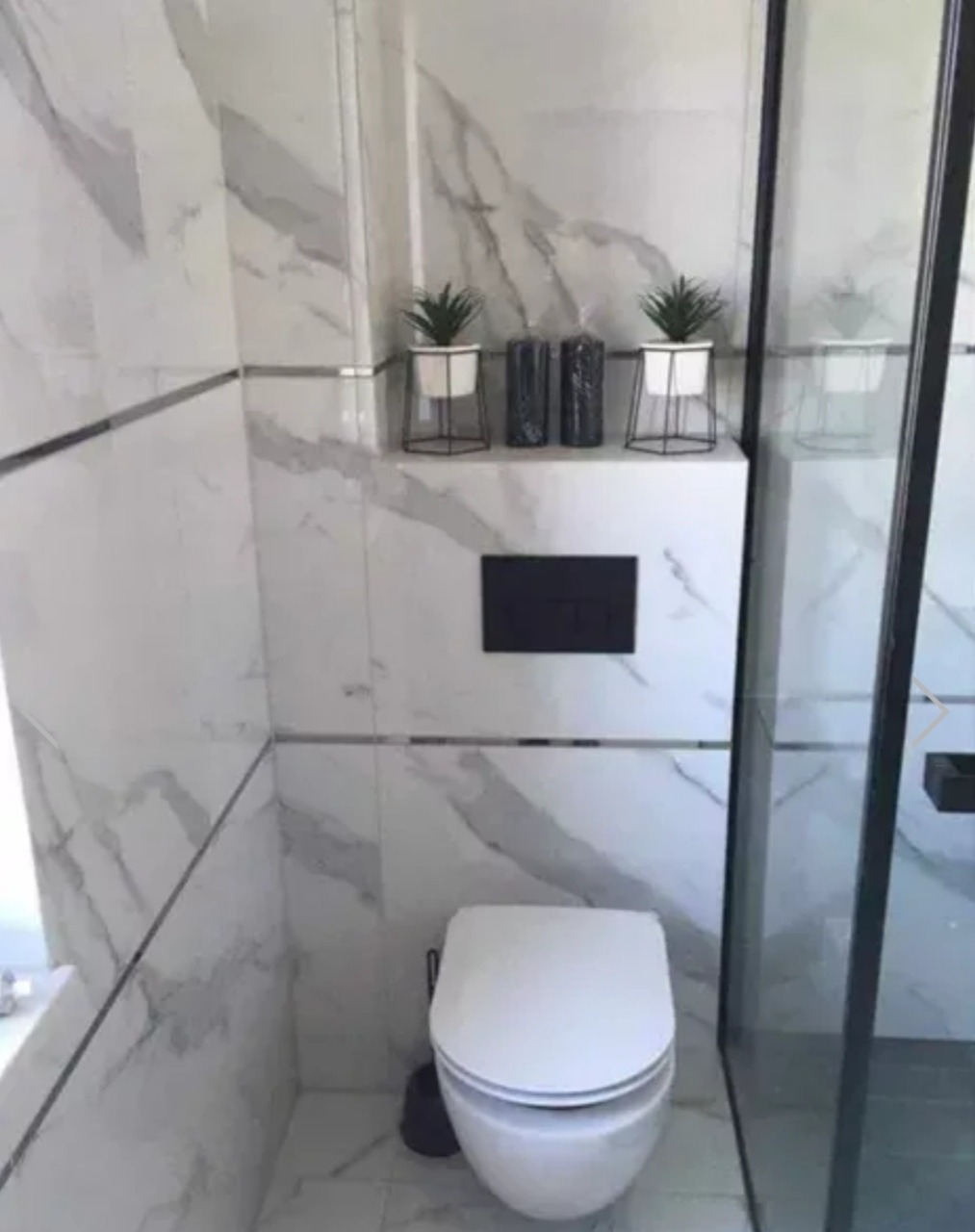 Nowoczesna toaleta podwieszana z marmurowymi płytkami na ścianach, minimalistycznymi dekoracjami roślinnymi i czarnymi akcentami, widoczna kabina prysznicowa.