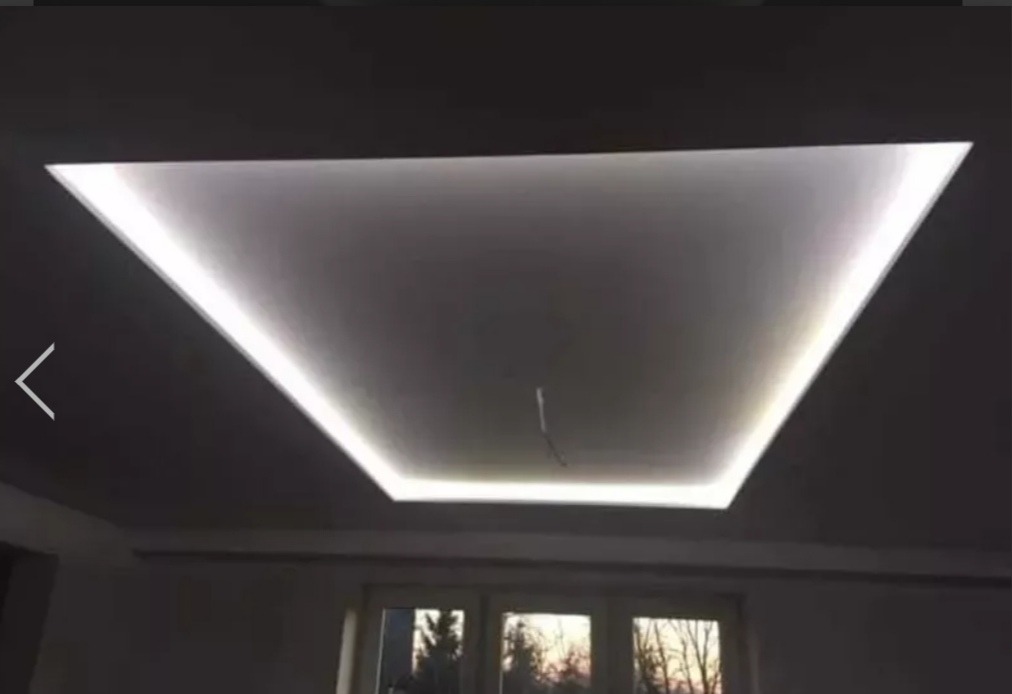 Nowoczesny sufit podwieszany z oświetleniem LED w kształcie trapezu, widok z dołu na tle okna.
