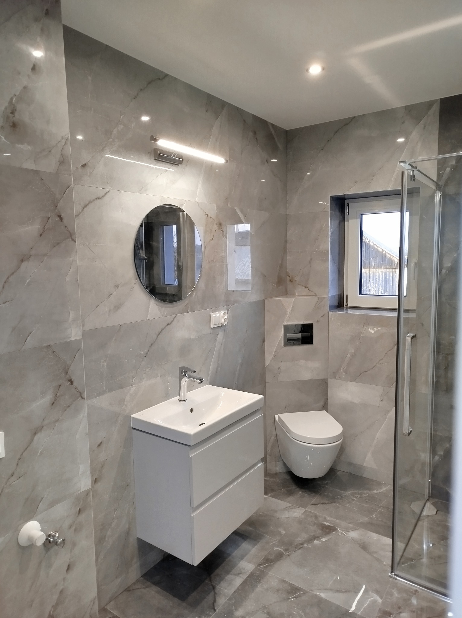 Nowoczesna łazienka wykończona płytkami imitującymi marmur, z wiszącą szafką z umywalką, okrągłym lustrem i kabiną prysznicową ze szklanymi drzwiami.