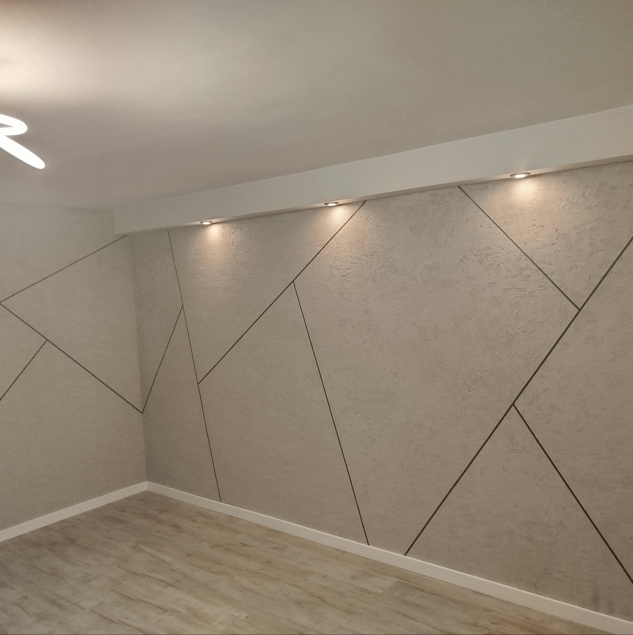 Ściana wewnętrzna z dekoracyjnym tynkiem strukturalnym i geometrycznym wzorem z ciemnych linii, oświetlona punktowymi lampami sufitowymi, z widoczną podłogą z paneli.