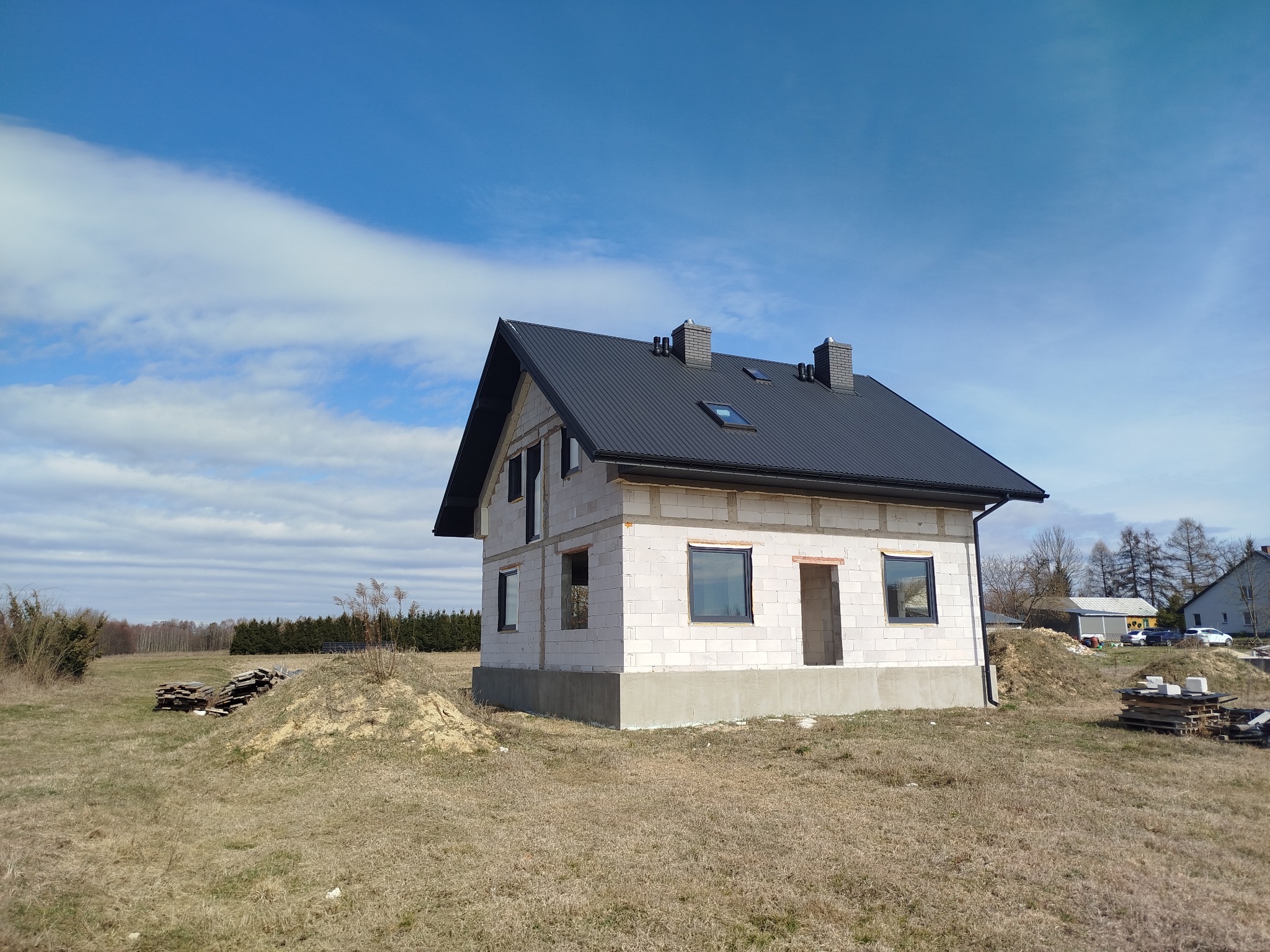 Niedokończony dwukondygnacyjny budynek mieszkalny z czarnym dachem i ceglanymi kominami, widoczne okna z czarnymi ramami, w tle błękitne niebo z chmurami i sterta drewnianych palet na trawiastym...
