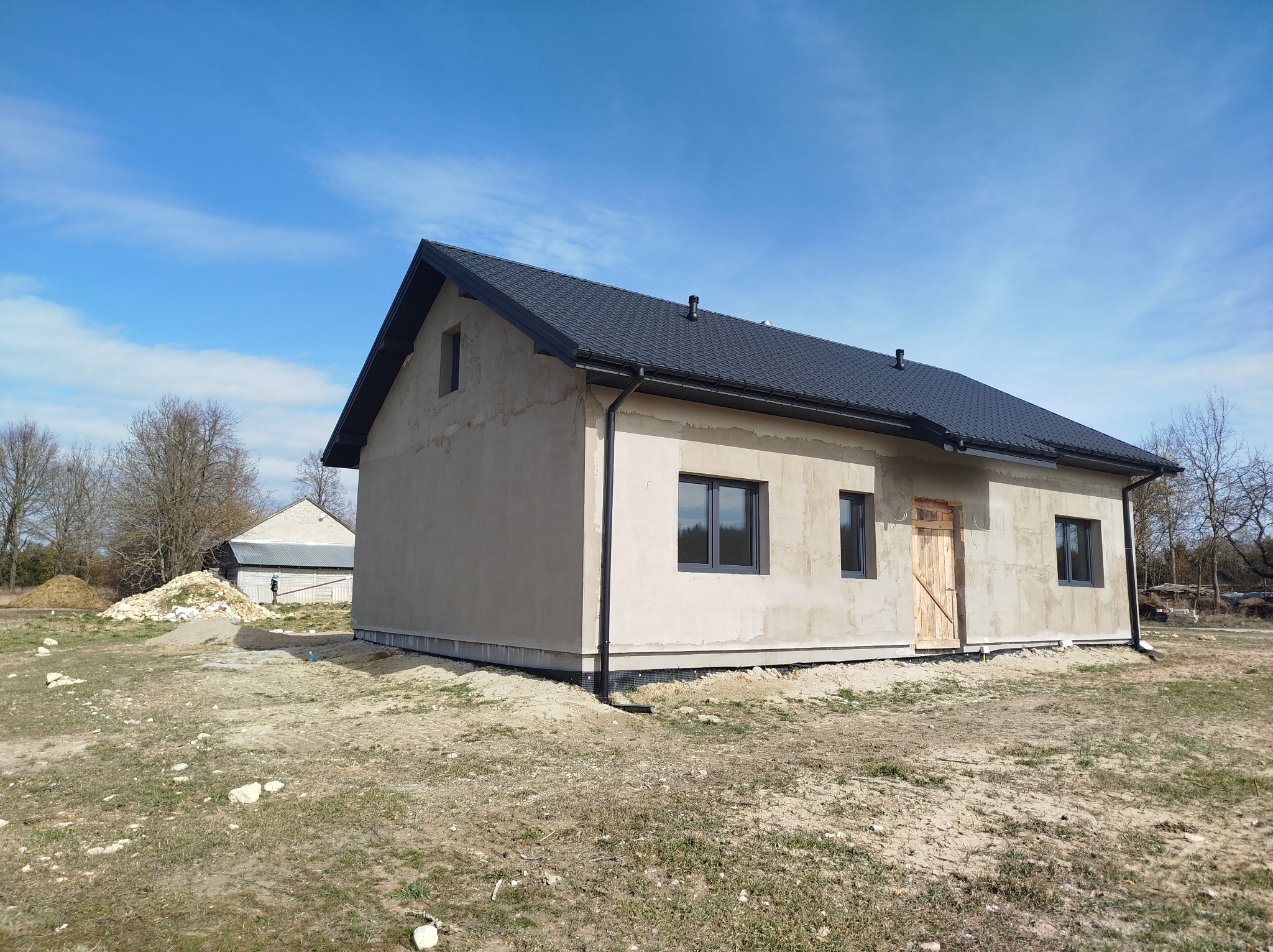 Surowy, otynkowany dom jednorodzinny z ciemną dachówką i szarą stolarką okienną, na działce z widoczną ziemią i trawą, w tle budynek gospodarczy i drzewa.