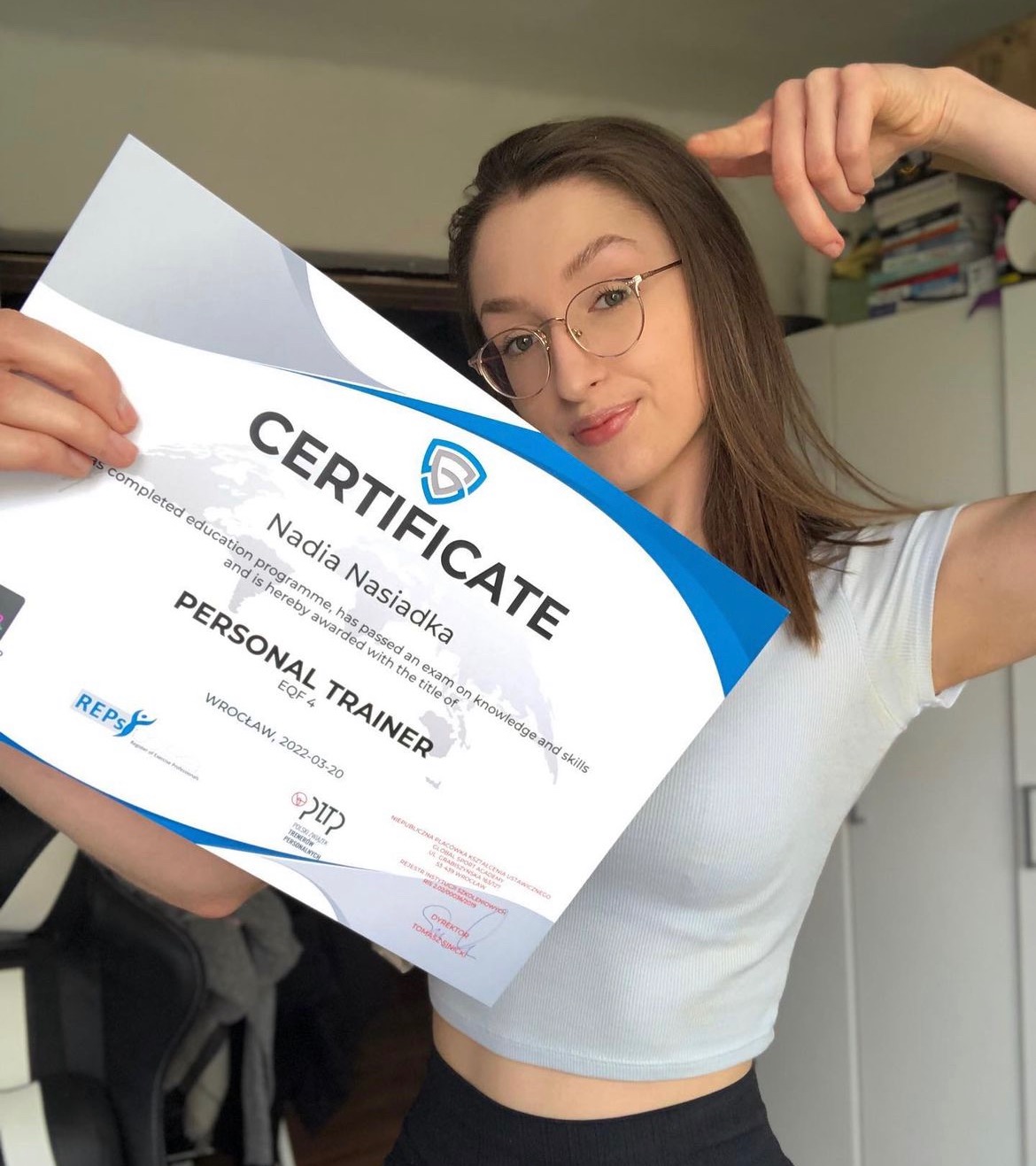 Uśmiechnięta kobieta w okularach prezentuje certyfikat ukończenia kursu trenera personalnego, trzymając go w dłoniach na tle wnętrza pokoju.