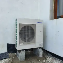 Biała jednostka zewnętrzna klimatyzacji marki Samsung, zamontowana na betonowych podstawach przy budynku w Stronie Śląskie, widoczne szare kamienie i fragment okna.