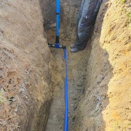Apt-Instal Paweł Andrełowicz - Wykop z ułożoną niebieską rurą i złączkami, obok stojący w wykopie pracownik w roboczych spodniach i butach.