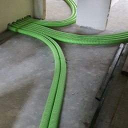 Apt-Instal Paweł Andrełowicz - Zielone, karbowane rury wentylacyjne ułożone na betonowej podłodze w surowym wnętrzu budynku, widoczne deski i stelaż krzesła w tle.