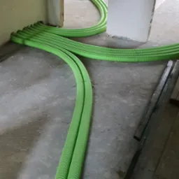 Zielone, karbowane rury wentylacyjne ułożone na betonowej podłodze w surowym wnętrzu budynku, widoczne deski i stelaż krzesła w tle.