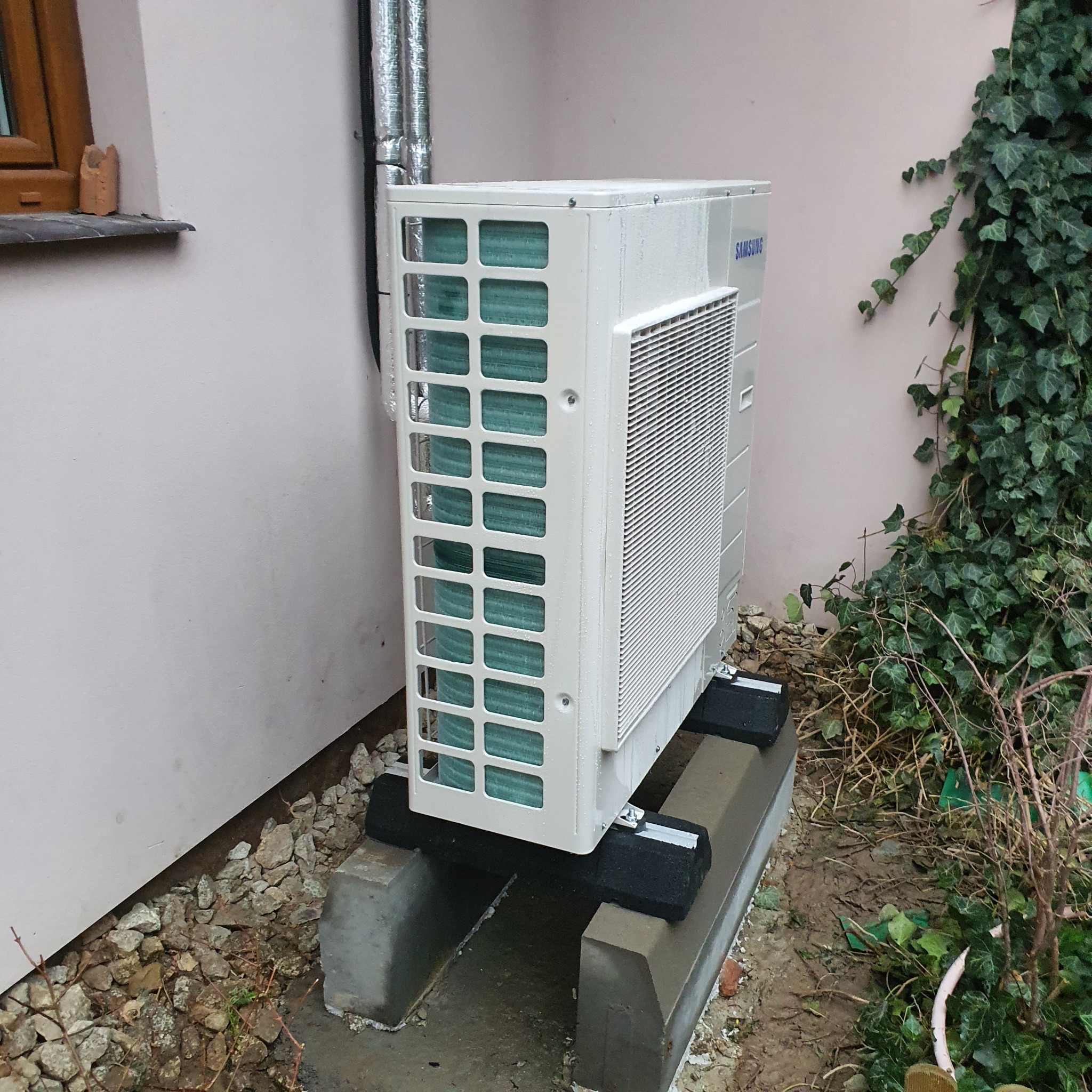 Zewnętrzna jednostka klimatyzacji Samsung zamontowana na betonowej podstawie z gumowymi podkładkami, obok ściany budynku z bluszczem.