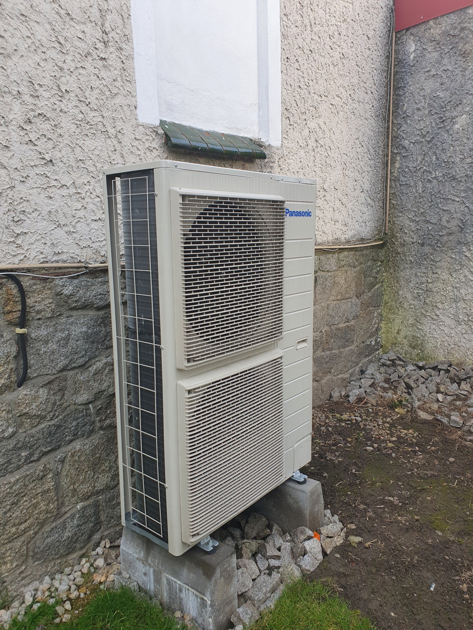 Zewnętrzna jednostka pompy ciepła Panasonic zamontowana na betonowych fundamentach przy nierównej, kamiennej ścianie budynku z widoczną instalacją elektryczną.