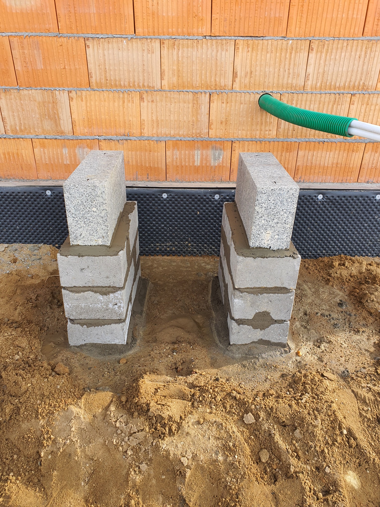 Wznoszenie fundamentów pod budynek z pustaków ceramicznych, widoczne dwa filary z bloczków betonowych z nakładkami z granitu, przyłącze rur w ścianie.