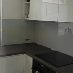 Szara szklana ściana nad blatem kuchennym w Grodzisku Mazowieckim, widoczne gniazdka elektryczne, okno z żaluzjami i fragment zlewu.