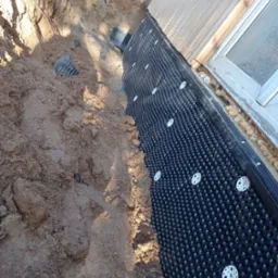 Czarna folia kubełkowa przymocowana do ściany fundamentowej za pomocą białych kołków, widoczny wykop z ziemią, okno w drewnianej ramie i pianka uszczelniająca.