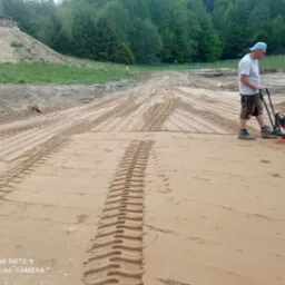 Pracownik w niebieskiej czapce zagęszcza piasek zagęszczarką wibracyjną na przygotowanym terenie budowy, widoczne ślady opon na piasku, w tle budynek w trakcie budowy i zalesiony teren.