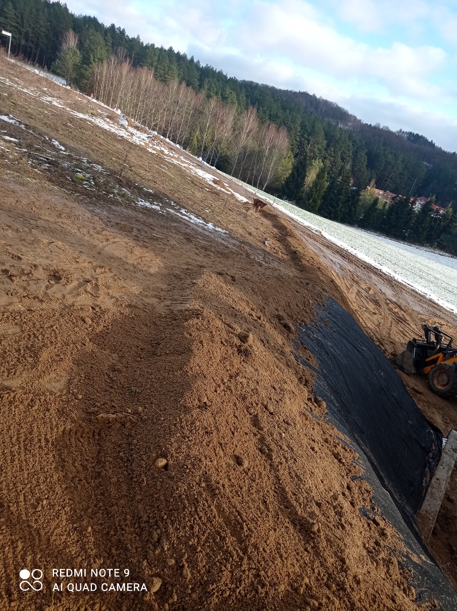 Przygotowanie terenu na zboczu: rozplantowana ziemia, czarna geowłóknina i mała ładowarka w prawym dolnym rogu, w tle las i częściowo zaśnieżone pole.