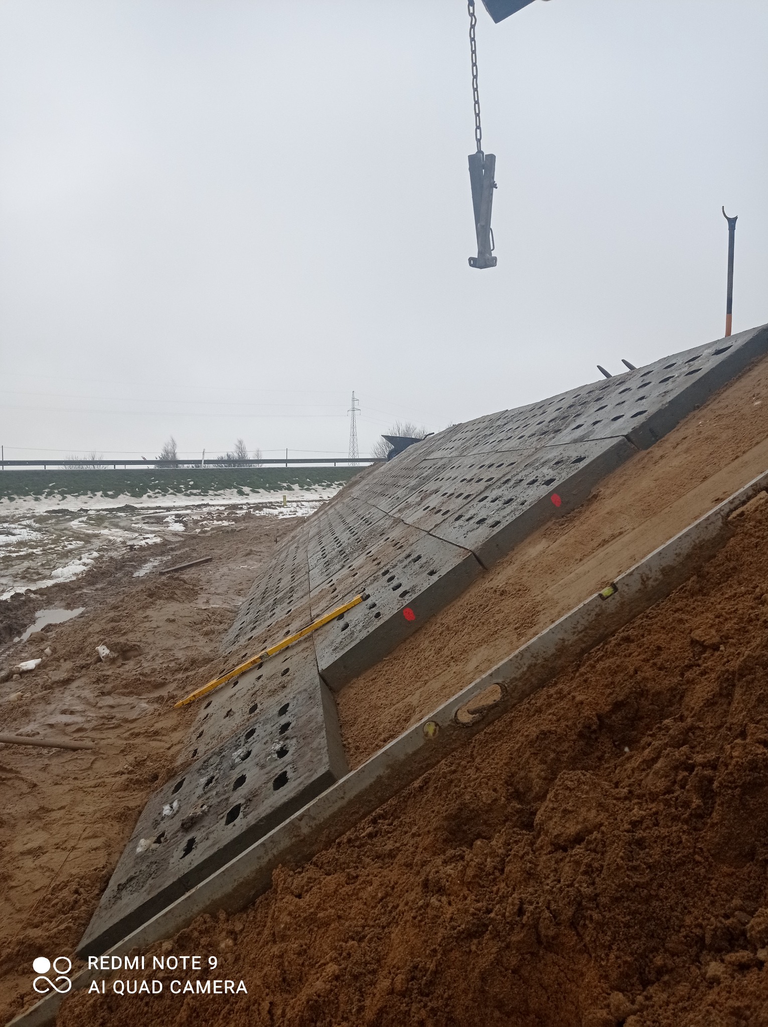 Budowa skarpy z prefabrykatów betonowych z otworami, poziomica przyłożona do krawędzi, ziemia wokół, pochmurne niebo.