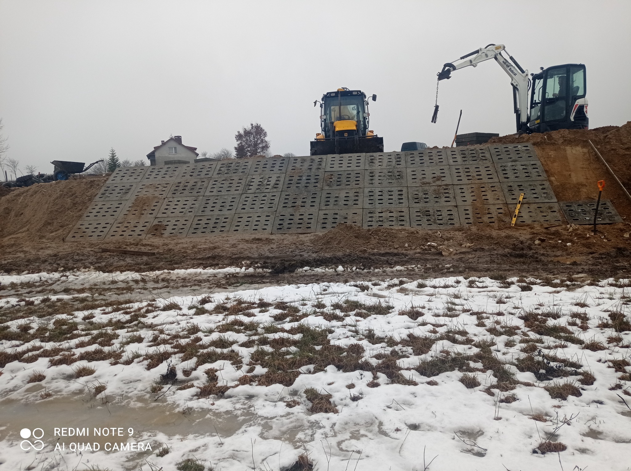 Wzmocnienie skarpy prefabrykowanymi elementami betonowymi, na górze koparka i traktor, w tle dom w zabudowie jednorodzinnej, zimowa sceneria z resztkami śniegu.