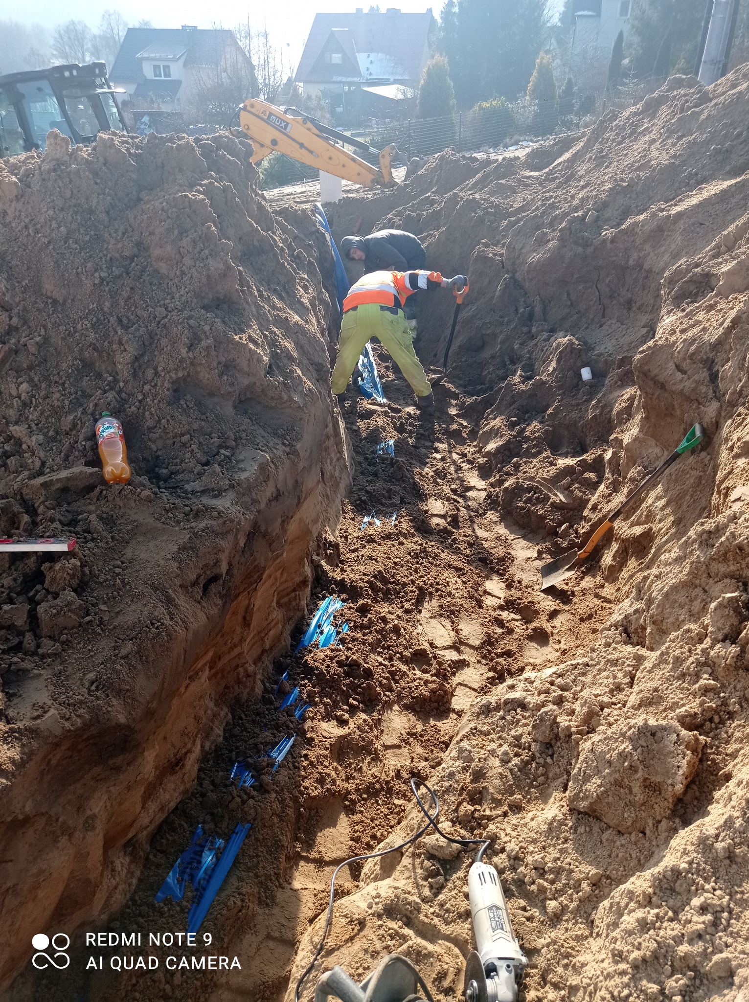 Głęboki wykop w ziemi z widocznymi niebieskimi rurami, pracownik w odblaskowej kamizelce kopie łopatą, w tle koparka, na krawędzi wykopu butelka napoju i poziomica, na dole szlifierka kątowa.