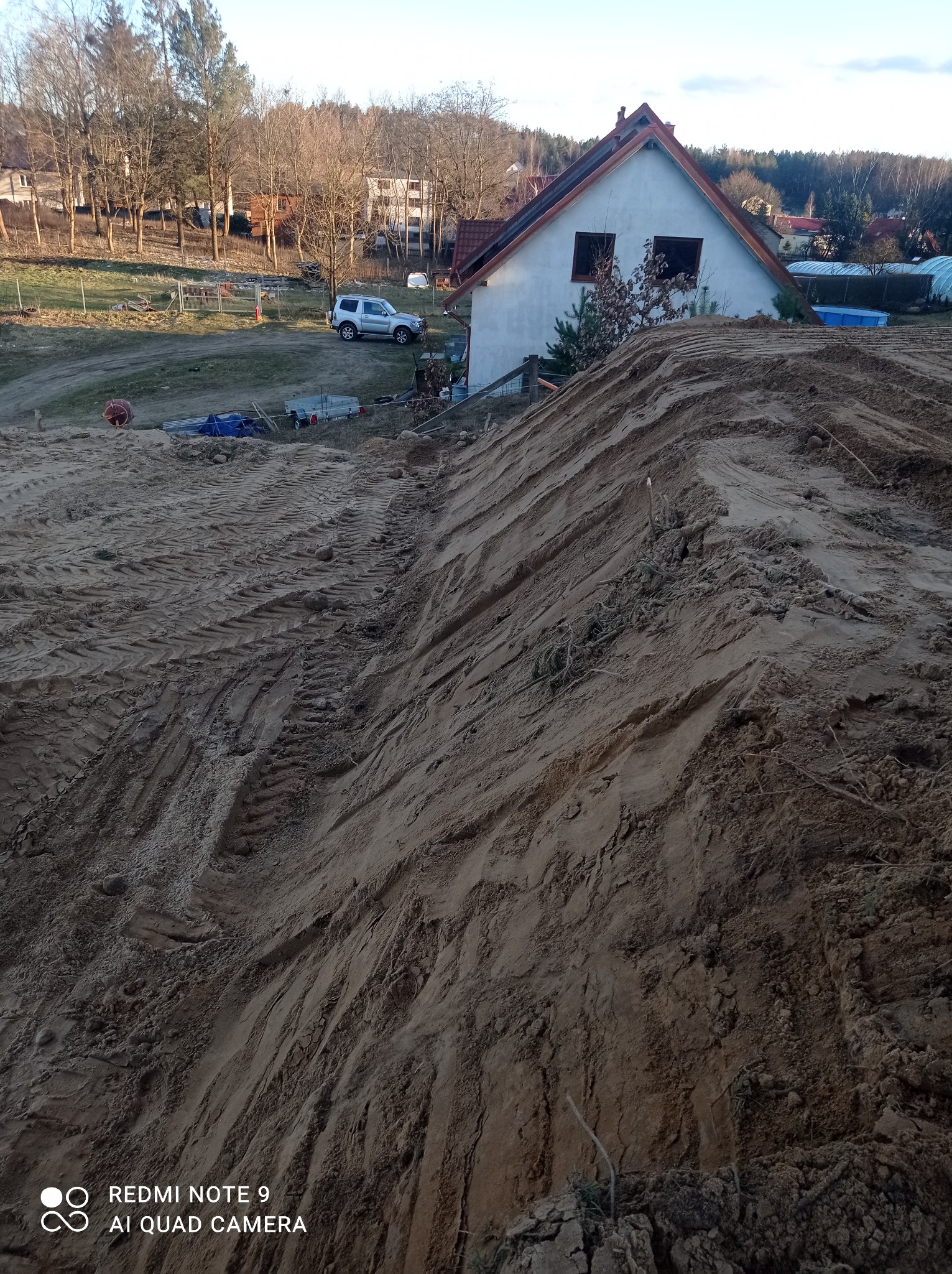 Kupa piasku z widocznymi śladami opon ciężkiego sprzętu budowlanego, w tle dom jednorodzinny z białymi ścianami, teren budowy w trakcie prac ziemnych.
