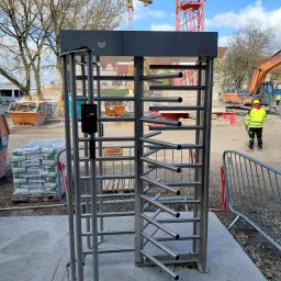 Luneco Sp. z o.o. - Metalowy kołowrót z czytnikiem kart na budowie, w tle żurawie i pracownik w odblaskowej kamizelce. System kontroli dostępu zintegrowany z alkomatem.