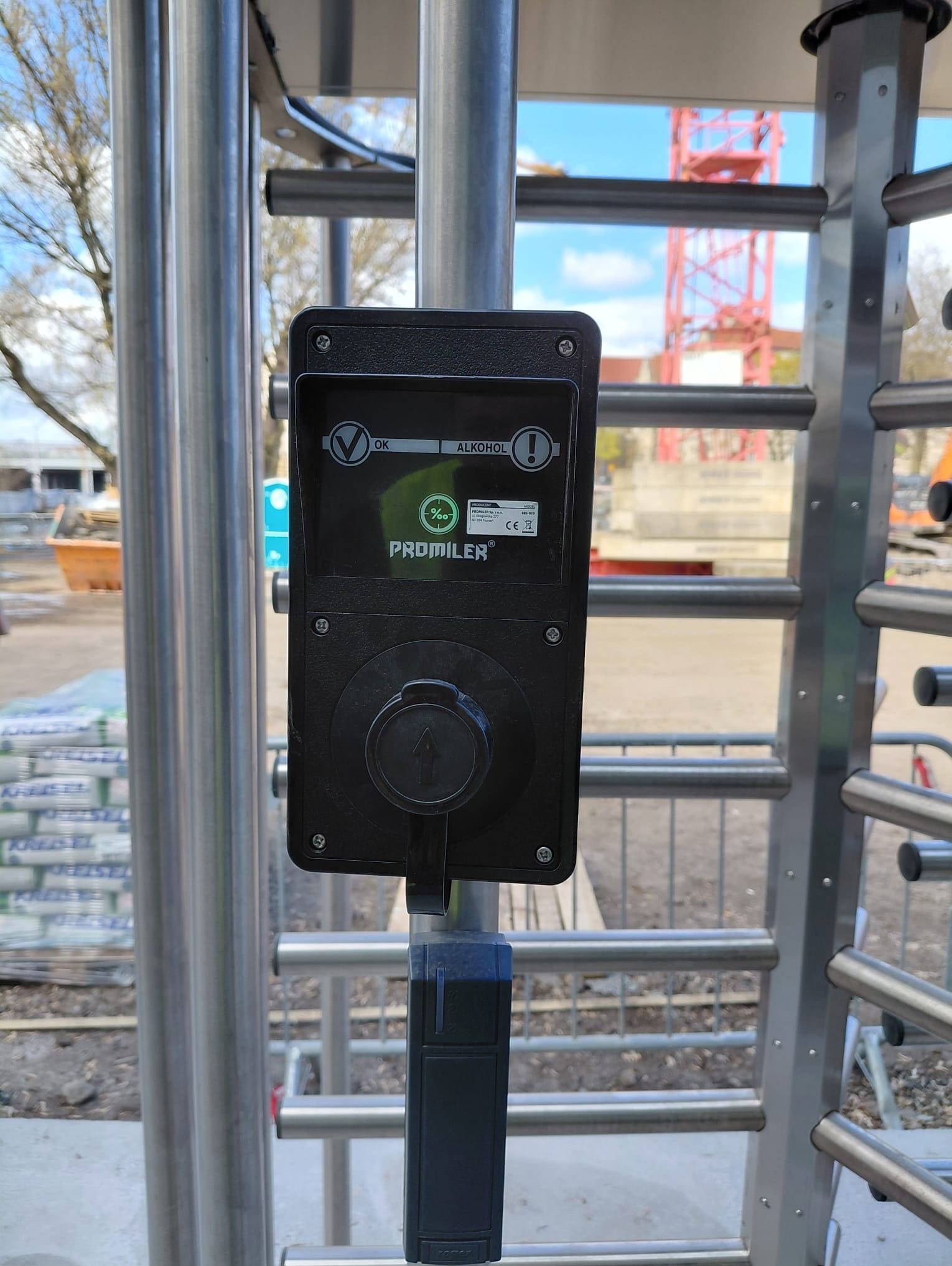 Zamontowany alkomat przy bramce obrotowej na budowie. Urządzenie marki Promiler z wyświetlaczem i pokrętłem. W tle widoczny żuraw budowlany i teren budowy.