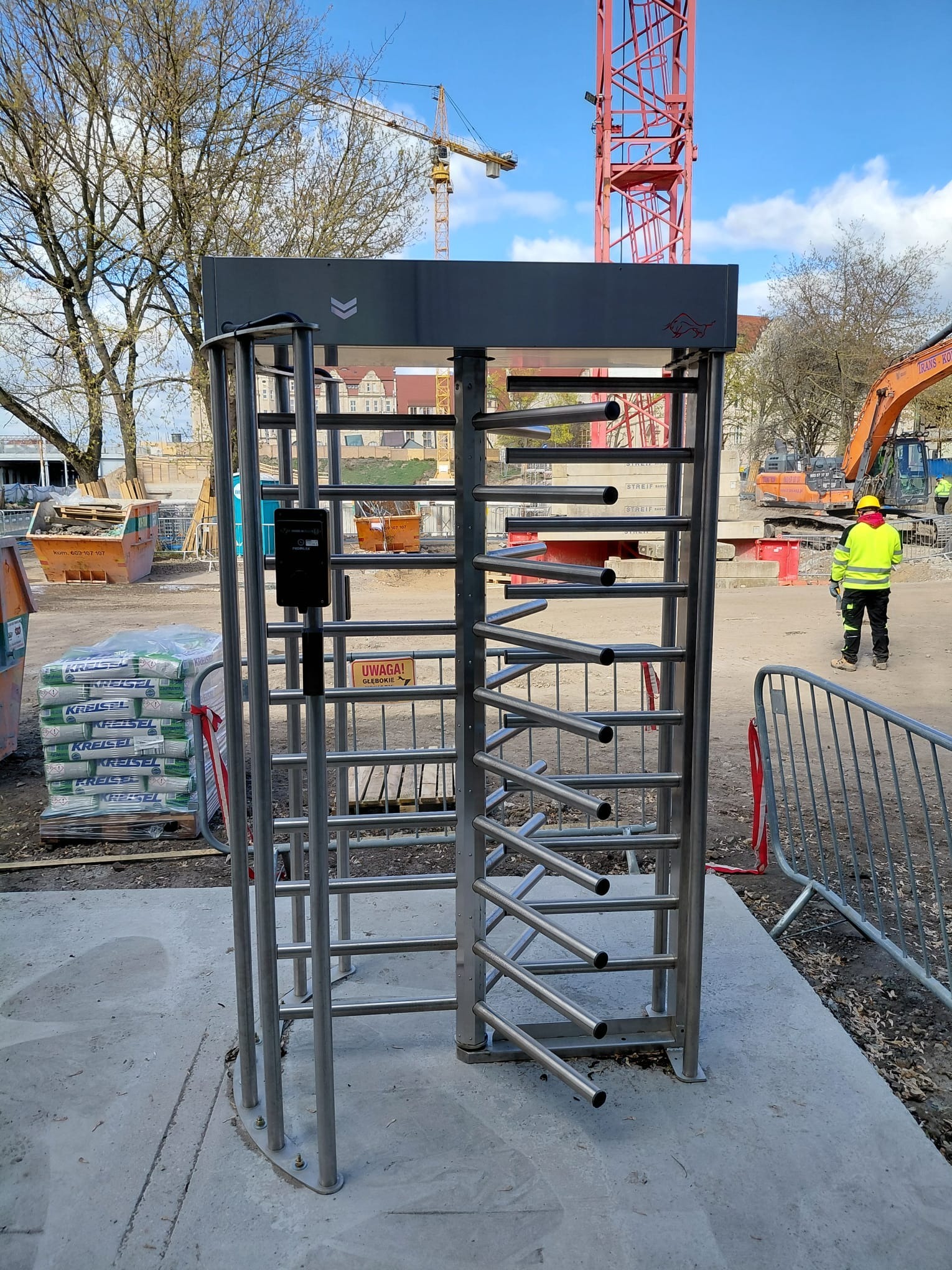 Metalowy kołowrót z czytnikiem kart na budowie, w tle żurawie i pracownik w odblaskowej kamizelce. System kontroli dostępu zintegrowany z alkomatem.