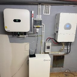 DREO SPÓŁKA Z OGRANICZONĄ ODPOWIEDZIALNOŚCIĄ - Instalacja z dwoma białymi falownikami i modułem bateryjnym Power Lab w szarej piwnicy z rurami grzewczymi pod sufitem.