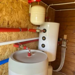 Dwa białe zbiorniki akumulacyjne ciepła różnej wielkości, połączone rurami w pomieszczeniu wyłożonym drewnem i płytami OSB, z widocznym manometrem na górze instalacji.