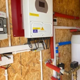 Instalacja fotowoltaiczna w pomieszczeniu technicznym: biały inwerter z żółtą naklejką ostrzegawczą, rozdzielnica elektryczna z napisem 'Główny wyłącznik AC', zbiornik na wodę z rurami w izolacji...