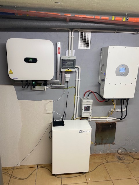 Instalacja z dwoma białymi falownikami i modułem bateryjnym Power Lab w szarej piwnicy z rurami grzewczymi pod sufitem.