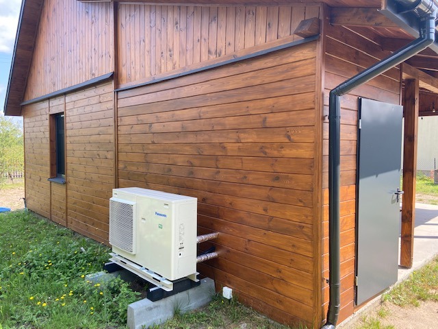 Zewnętrzna jednostka klimatyzacji Panasonic zamontowana na betonowych bloczkach przy drewnianym budynku z ciemnobrązową elewacją, widoczna rynna i szara metalowa furtka.