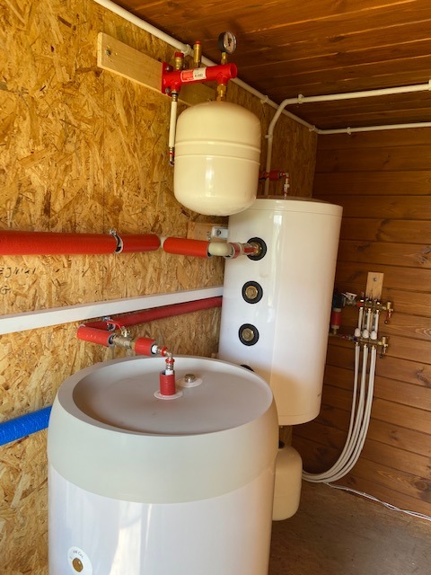 Dwa białe zbiorniki akumulacyjne ciepła różnej wielkości, połączone rurami w pomieszczeniu wyłożonym drewnem i płytami OSB, z widocznym manometrem na górze instalacji.