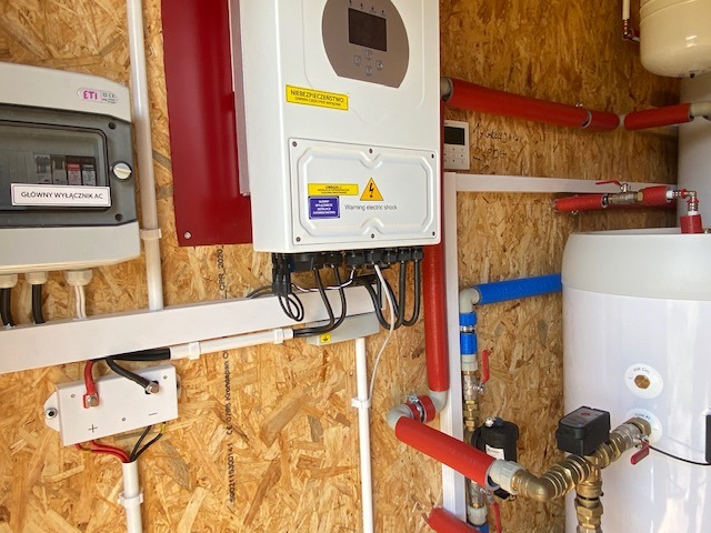 Instalacja fotowoltaiczna w pomieszczeniu technicznym: biały inwerter z żółtą naklejką ostrzegawczą, rozdzielnica elektryczna z napisem 'Główny wyłącznik AC', zbiornik na wodę z rurami w izolacji...