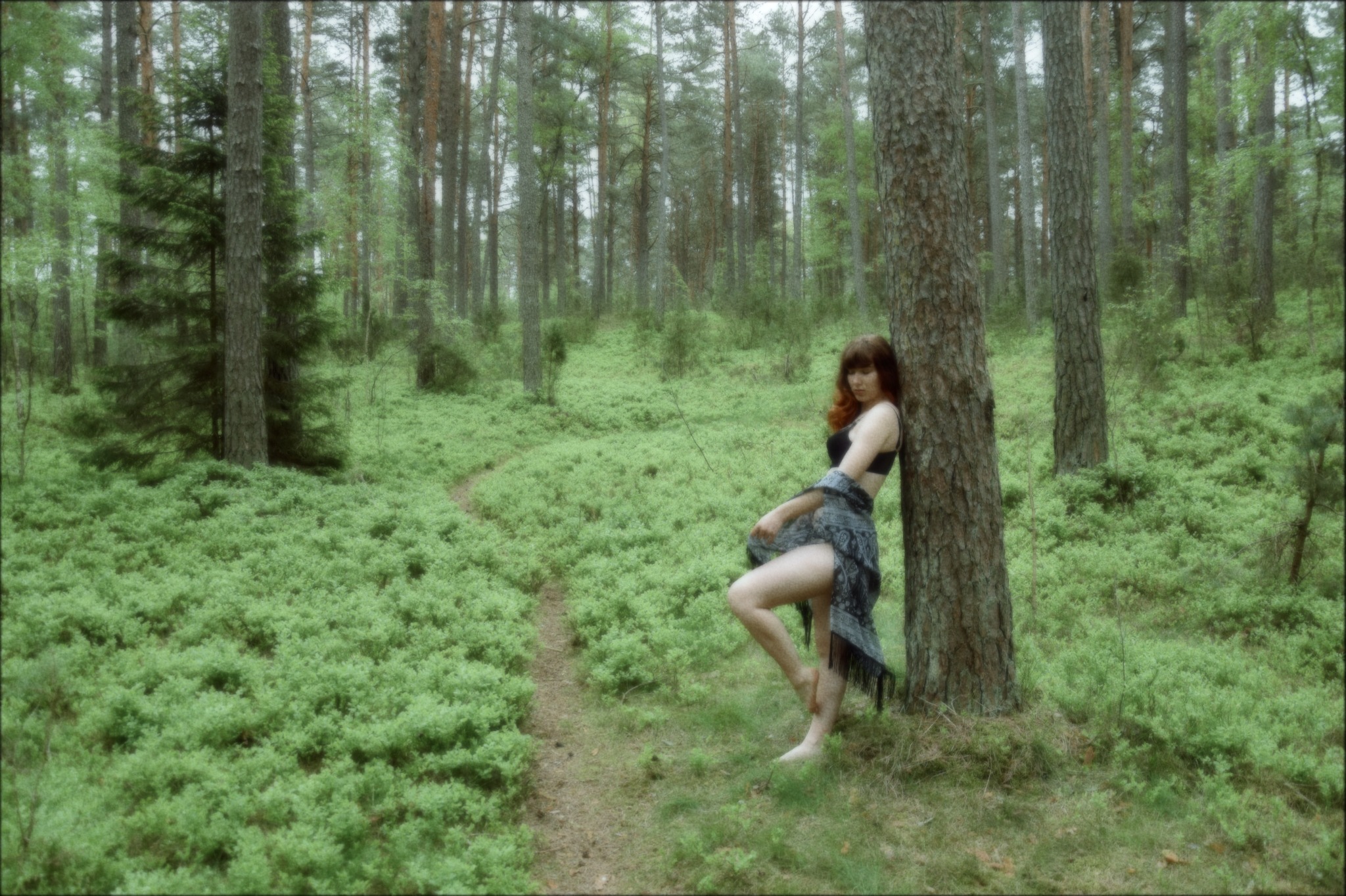 Kobieta z rudymi włosami opiera się o drzewo w lesie, ubrana w czarny top i szarą chustę z frędzlami, ścieżka z zielonych roślin w tle.