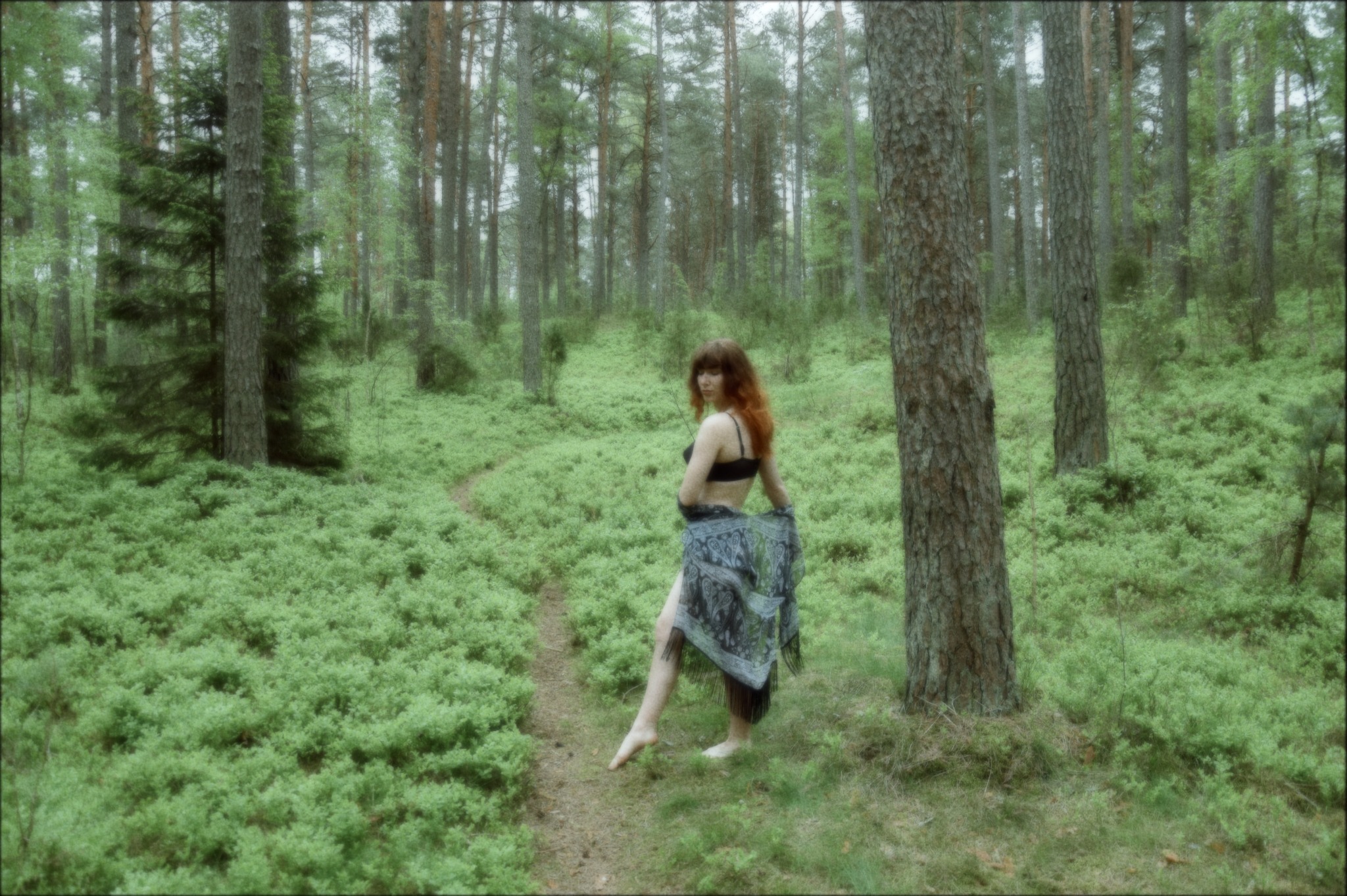 Ruda kobieta w czarnym biustonoszu i wzorzystej chuście pozuje boso w zalesionym krajobrazie, odwrócona plecami do obiektywu, z delikatnym uśmiechem.